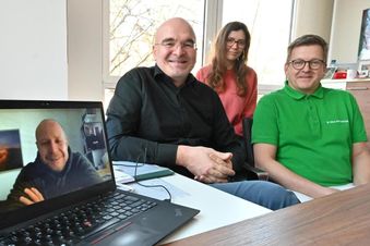 Drei Personen in einem Büro nehmen an einem Videomeeting teil, eine vierte Person ist per Laptop zugeschaltet.