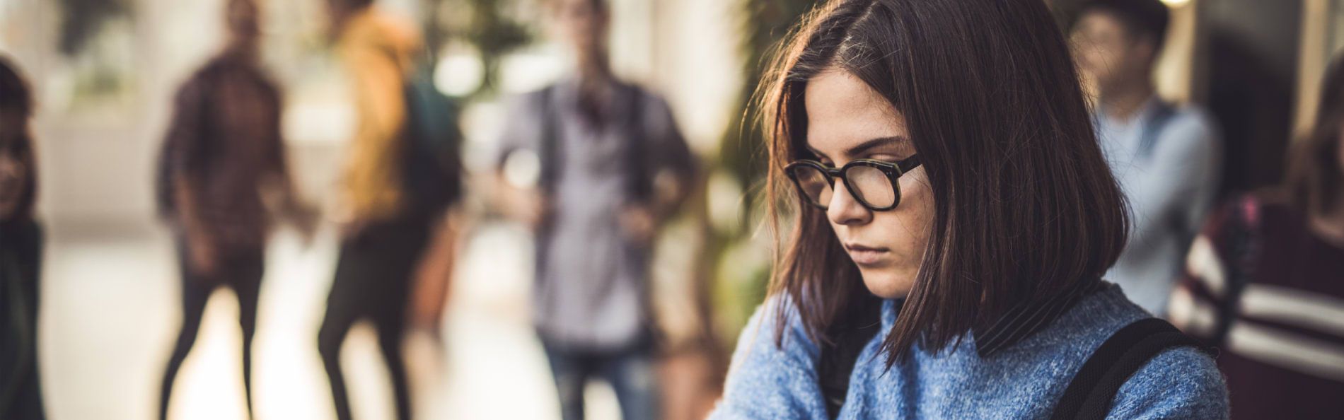 Eine Schülerin mit Brille steht mit gesenktem Blick abseits einer Schülergruppe 