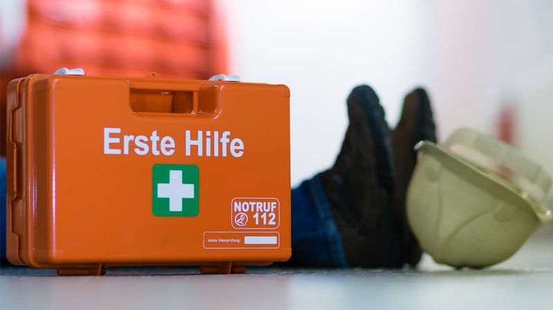 Ein Erste-Hilfe-Kasten steht auf dem Boden, neben einer liegenden Person und einem Arbeitshelm.