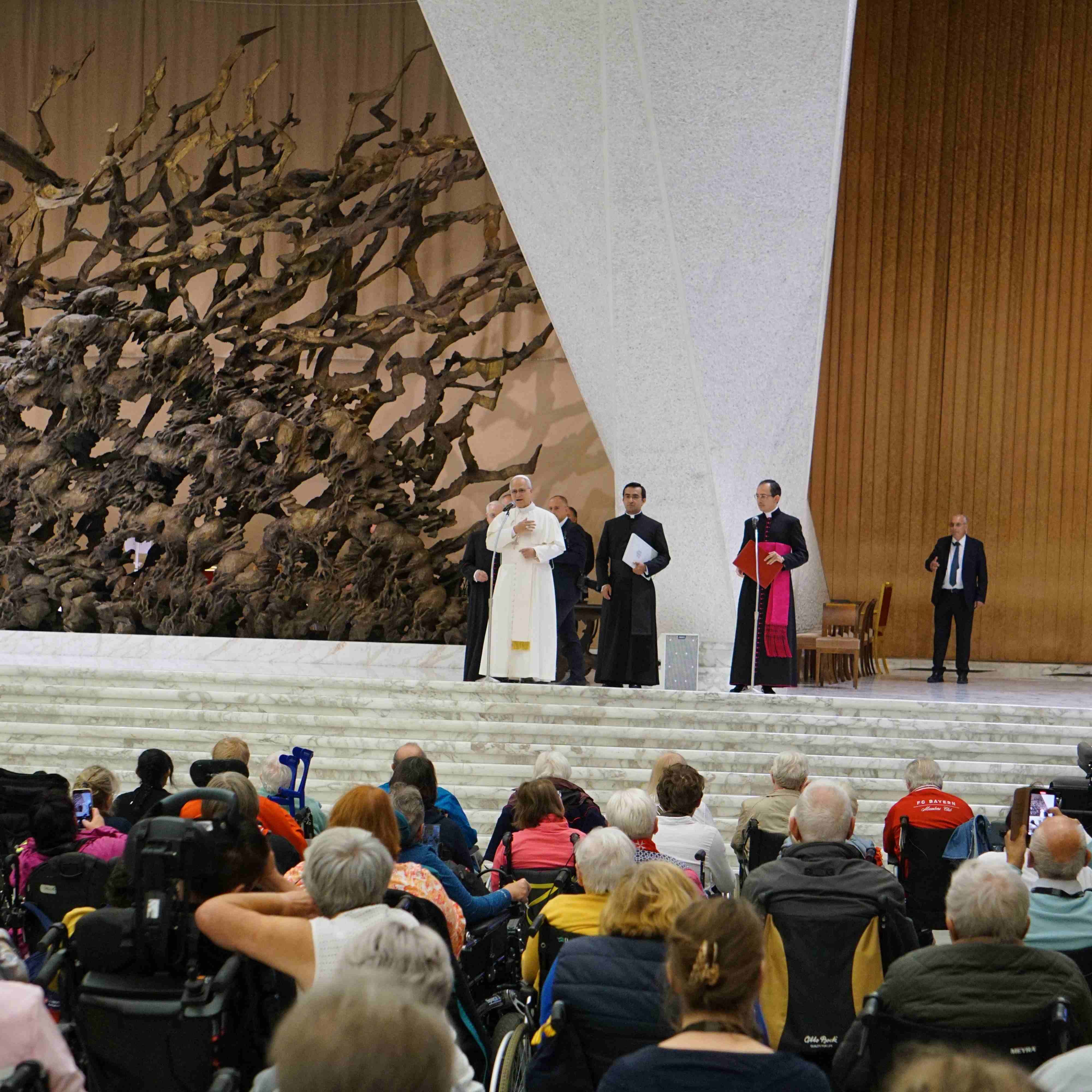 Vier Personen in religiöser Kleidung, in der Mitte Papst Leo XIV.,stehen in der Audienzhalle auf einer Treppe vor einer großen, abstrakten Skulptur und einem Holzpaneel, Publikum sitzt davor.