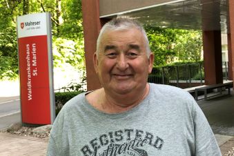 Älterer Mann in grauem T-Shirt steht vor dem Eingang einer Klinik mit rotem Hinweis-schild.