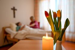 Symbolbild, das im Vordergrund einen Blumenstrauß und im Hintergrund einen sterbenden, begleiteten Menschen im Bett zeigt. (Foto: Malteser)