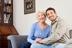 Eine ältere Dame mit Hausnotruf-Knopf sitzt mit ihrem Sohn auf dem Sofa und lacht