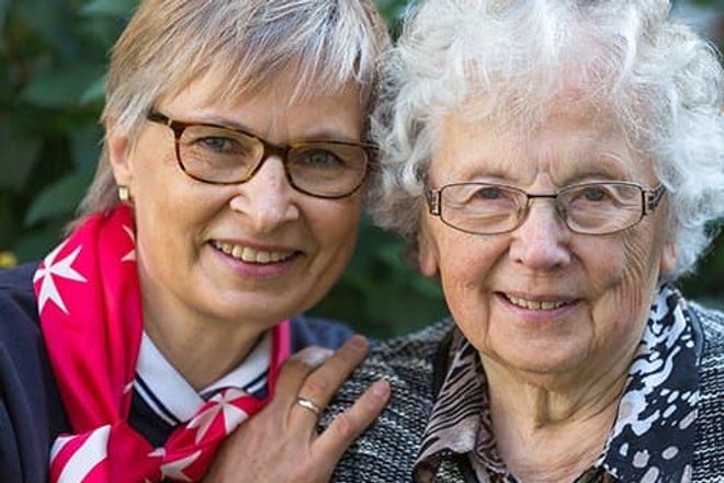 Malteser Mitarbeiterin legt Arm um ältere Frau und beide lachen in die Kamera.