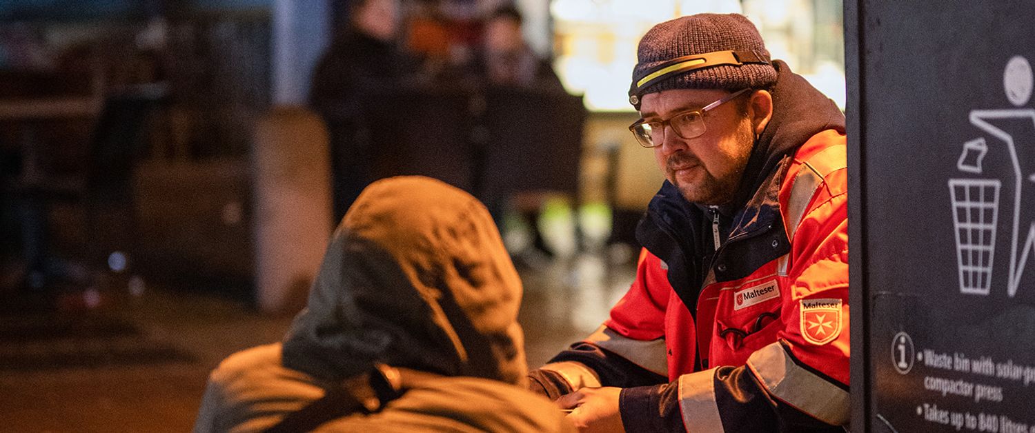 Ein Malteser kniet vor einem Obdachlosen auf der Straße und spricht mit ihm.