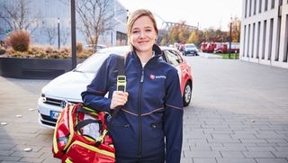 Mitarbeiterin der Malteser steht mit Erste-Hilfe-Koffer lächelnd vor einem Auto der Malteser.