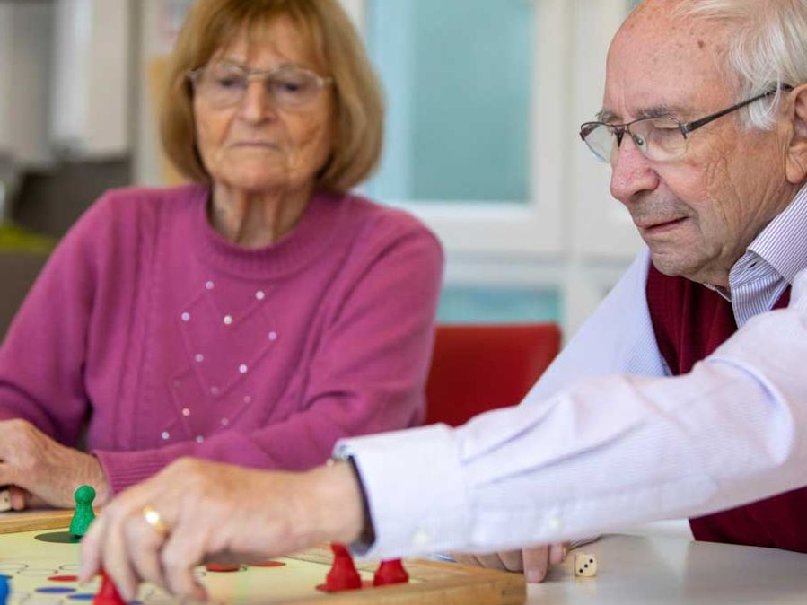 Ein älterer Mann greift zu einer Spielfigur eines Brettspiels, neben ihm sitzt eine ältere Frau.