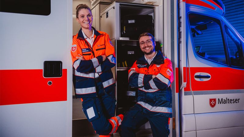 Eine junge Frau und ein junger Mann an einem Rettungswagen der Malteser