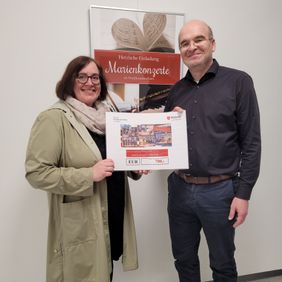 Frau und Mann halten gemeinsam einen Spendenscheck, im Hintergrund ein Plakat zu einer Konzertreihe.