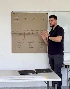 Mann in dunkelblauem Malteser Outfit steht an einem Plakat und erklärt