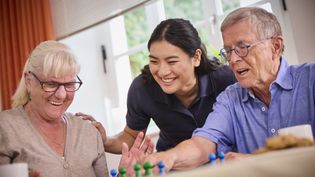 Drei Personen sitzen gemeinsam an einem Tisch vor einem Fenster und spielen ein Brettspiel mit bunten Spielfiguren. Eine Person im Hintergrund beugt sich unterstützend über die Spielenden, während Tassen und Spielmaterial auf dem Tisch stehen.