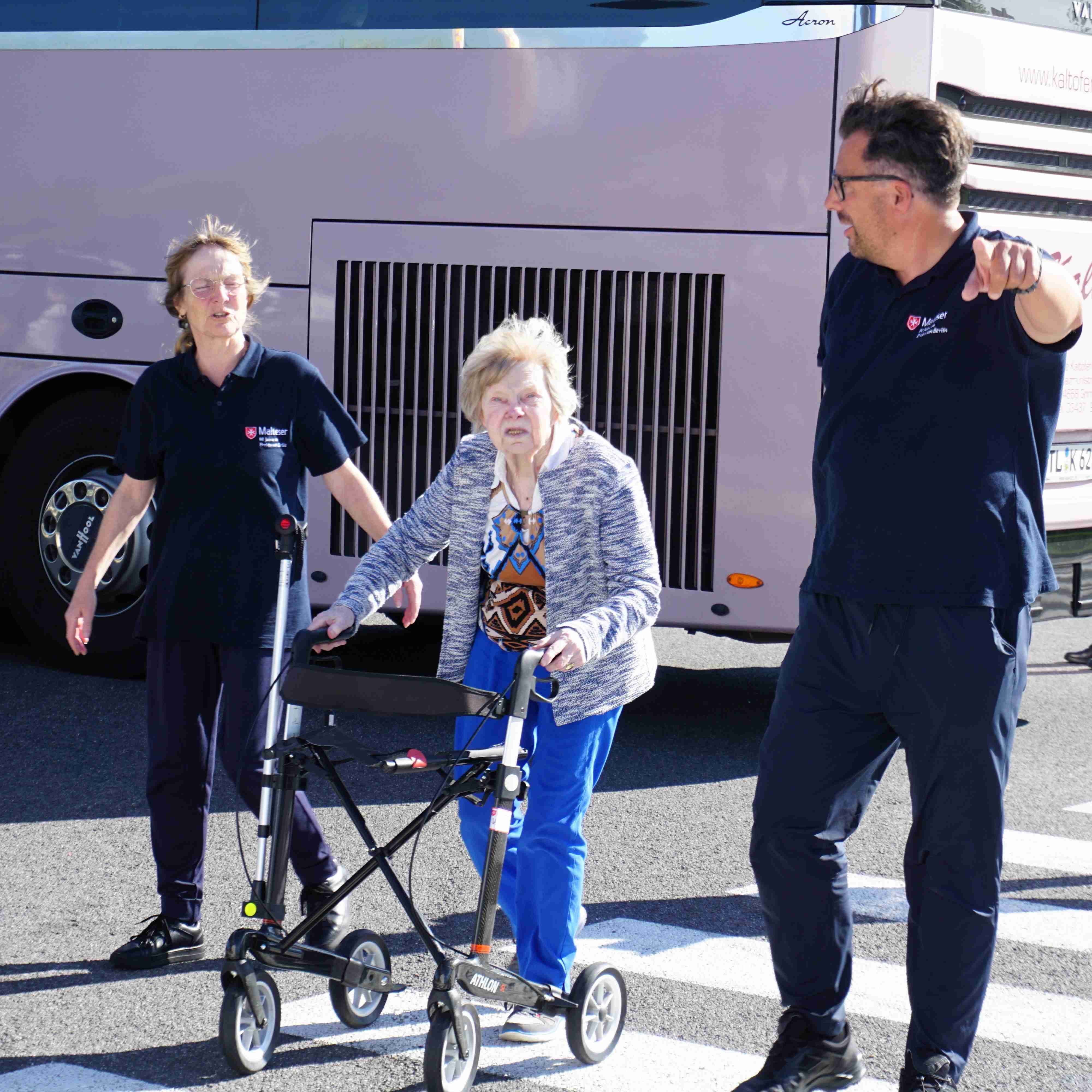 Ältere Frau mit Rollator steht auf Straße vor Reisebus, begleitet von zwei Personen in dunkler Malteser Kleidung, eine zeigt nach rechts.