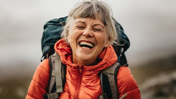 Ältere Dame mit oranger Jacke und Rucksack lacht zufrieden.