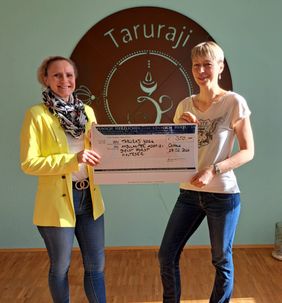 Zwei Personen stehen in einem Raum vor einer türkisfarbenen Wand mit dem großen runden Logo „Taruraji“. Beide halten gemeinsam einen großen Spendenscheck in den Händen. Auf dem Scheck ist eine Spende an den ambulanten Hospizdienst der Malteser in Forst vermerkt. Das Licht fällt warm durch den Raum und hebt Scheck und Logo deutlich hervor.