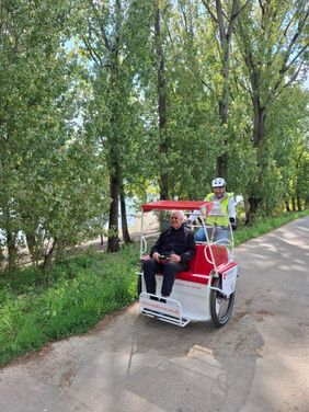 Ein Ehrenamtlicher fährt einen Mann in einer Fahrradrikscha am Rheinufer