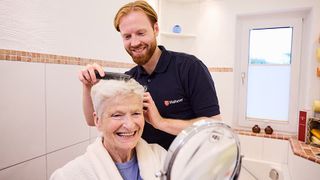 Ein Malteser Mitarbeiter kämmt einer älteren Frau in ihrem Badezimmer die Haare.