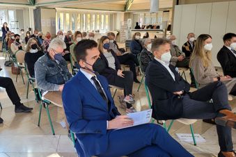 Personen sitzen mit Abstand und teils mit Maske bei einer Veranstaltung in einem Saal.