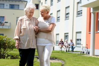 Malteser-Mitarbeiterin führt eine ältere Dame vor einem Pflegeheim