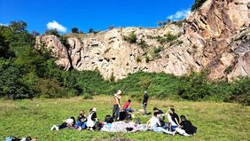 Menschen sitzen auf Decken auf einer Wiese. Im Hintergrund ist eine Felswand zu sehen.