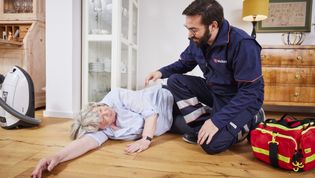 Ein Mann in Malteser Dienstbekleidung kniet neben einer gestürzten älteren Frau mit Hausnotrufknopf