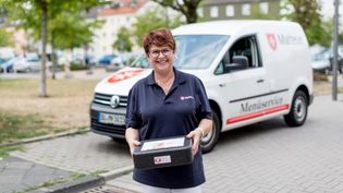Mitarbeiterin des Malteser Menueservice trägt eine Warmhaltebox und lächelt in die Kamera.