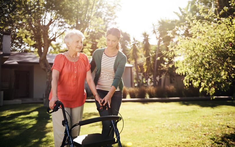 Frau hilft älterer Frau mit Rollator beim Gehen.