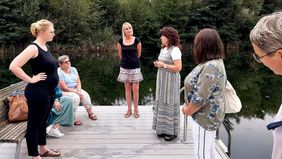 Frauen stehen auf einem Steg an einem See. Eine Frau spricht. Die anderen hören zu.