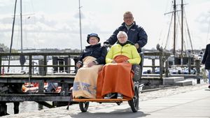 Zwei ältere Frauen sitzen nebeneinander in einem dreirädrigen Rollstuhl-Fahrrad und sind mit Decken zugedeckt, während ein Mann sie von hinten schiebt. Im Hintergrund ist ein Hafen mit Segelbooten und Stegen zu sehen, und die Gruppe bewegt sich entlang einer gepflasterten Uferpromenade.