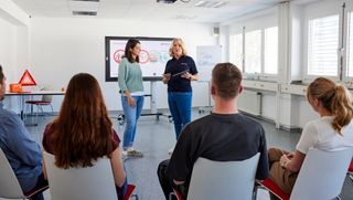 Ausbilderin der Ersten-Hilfe hält ein Tablet in der Hand und steht mit einer Kursteilnehmenden vor den anderen Teilnehmern.