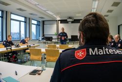 Ein Mann von hinten mit Malteser Jacke sitzt in einem Tagungsraum.