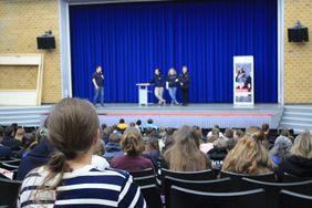 Begrüßungssituation während des Fachtags. Leitungen des Fachtags stehen auf der Bühne und begrüßen das Publikum, welches auf Stühlen vor ihnen sitzt. 