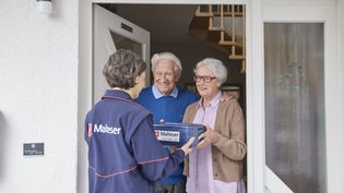Ein älterer Mann und eine ältere Dame stehen im Eingangsbereich ihres Hauses in der geöffneten Haustür. Eine Malteser-Mitarbeiterin steht den beiden gegenüber. Sie überreicht ihnen eine Essensbox. Sie nehmen diese an. 
