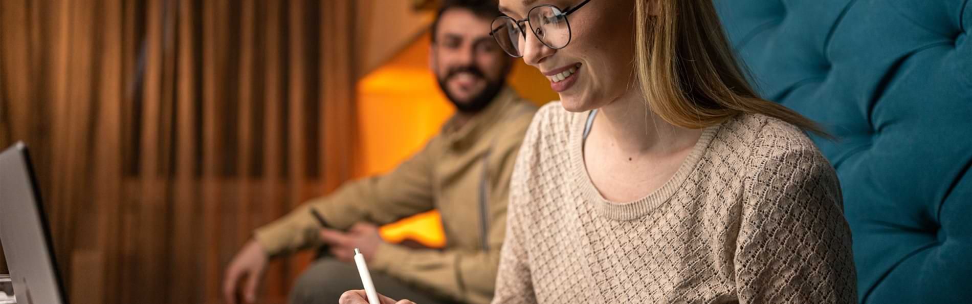 Ein lächelndes junges Paar auf einem Sofa. Die Frau im Vordergrund notiert etwas in einem Notizbuch.