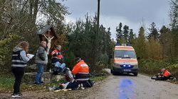 An einem Waldweg steht ein Krankenwagen, Menschen liegen auf dem Boden, Einsatzkräfte knien daneben, am Rand steht eine Beobachterin und schreibt