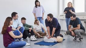 Ein Malteser Ausbilder demonstriert an einer auf dem Boden liegenden Übungspuppe die Herzdruckmassage, um ihn herum sitzen und stehen Kursteilnehmende
