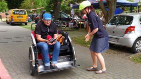 Ein Mann mit einem Mikrofon sitzt in einer Fahrradrikscha. Daneben steht eine Frau.