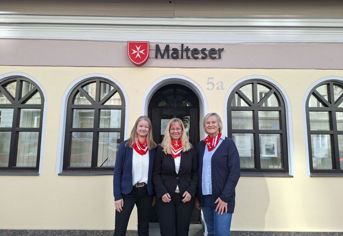 drei lachende Frauen stehen vor einem gelben Gebäude mit der Aufschrift Malteser und dem Malteser Logo