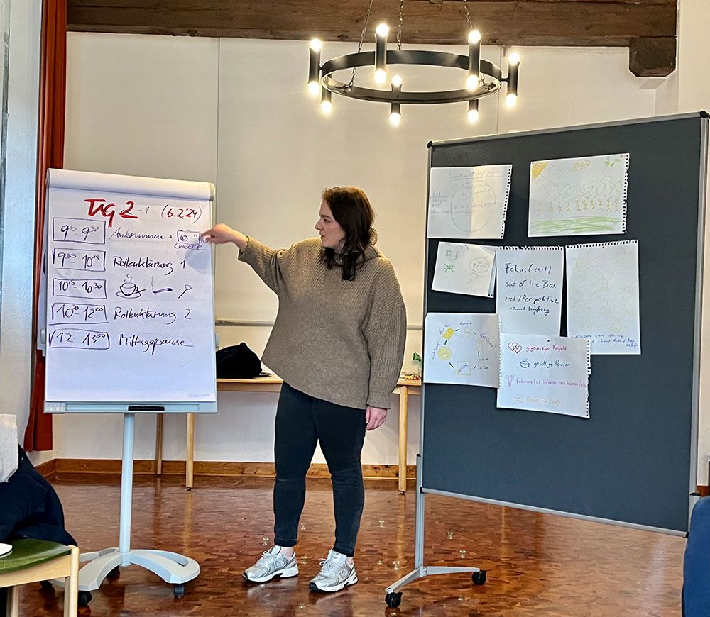 Clara Hüttenbrink-Marku steht zwischem einem Flipchart und einem Präsentationsboard. Auf dem Flipchart ist ein Tagesablauf mit Uhrzeiten und Programmpunkten wie ‚Begrüßung‘, ‚Reflexionsphase‘ und ‚Pause‘ notiert. Das Präsentationsboard daneben ist mit mehreren Blättern mit Zeichnungen und Notizen bestückt. Der Seminarraum hat einen Holzboden und einen Kronleuchter an der Decke.