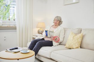 Kundin des Malteser Hausnotrufs trägt den Notfallknopf als Notrufarmband und ließt eine Zeitschrift auf dem Sofa.