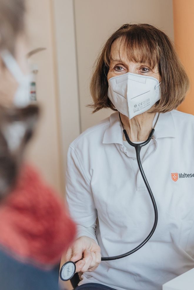 Frau Dr. Anne Rauhut mit einer Maske sitzt mit dem Gesicht frontal zu einem Patienten, den man im Vordergrund erkennen kann. Sie hält ein Stethoskop in der Hand. 