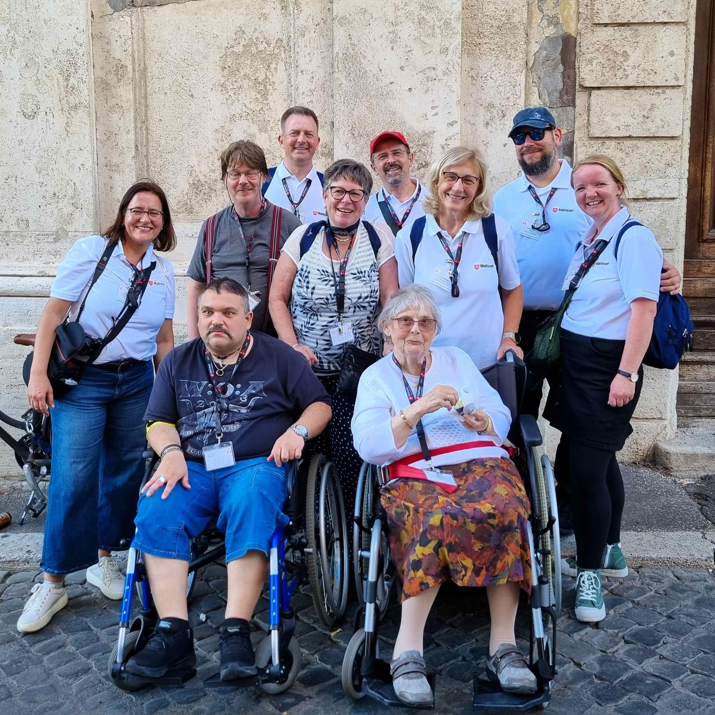 Gruppe von Menschen, darunter zwei Personen im Rollstuhl, vor einer Steinmauer im Freien, alle tragen lässige Kleidung.