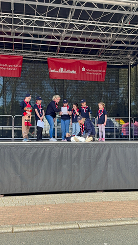 acht Kinder der Malteser Jugend Oberhausen sind auf der Bühne, ein Kind spricht in das Mikrofon, das ihm die Gruppenleiterin vorhält, ein Mädchen demonstriert auf Anweisung die stabile Seitenlage an einem Jungen