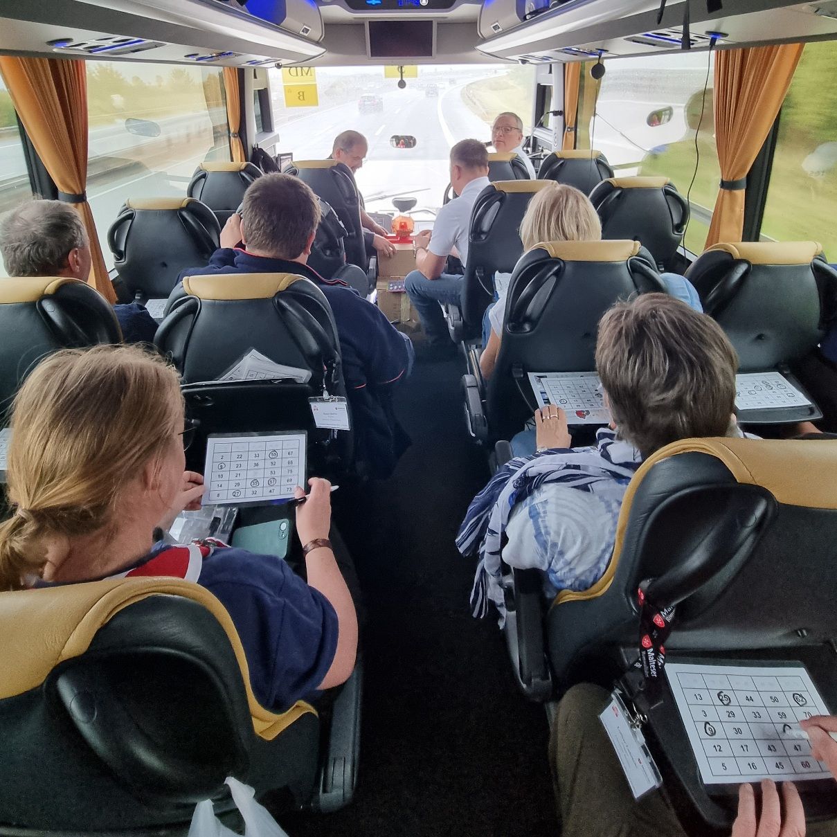 Innenraum eines Busses mit Fahrgästen, die auf ihren Sitzen sitzen und teilweise eine Zeitschrift halten.