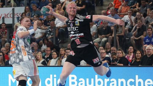 Handballspieler im Sprungwurf mit Ball, ein Gegenspieler verteidigt, weitere Personen im Hintergrund.