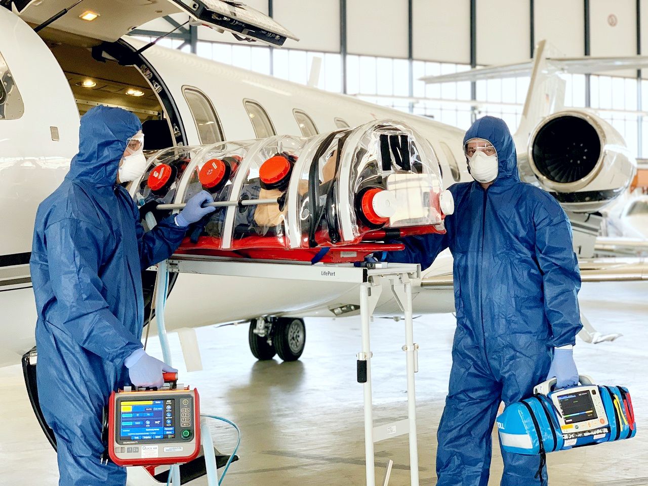 Zwei Männer in Schutzkleidung laden eine Bahre aus einem Flugzeug.