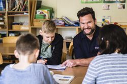 Mehrere Kinder sitzen gemeinsam an einem Tisch in einem Klassenraum, während ein lächelnder Erwachsener sie bei einer Schreibaufgabe unterstützt. Im Hintergrund sind Regale mit Büchern und Unterrichtsmaterialien zu sehen.
