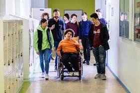 In einem Schulgang ist eine Gruppe von Schülerinnen und Schülern auf dem Weg ins Klassenzimmer. Inmitten der Gruppe ein junger Mann im Rollstuhl, der von einem Malteser Helfer begleitet wird. und in Begleitung eines Malteser helfers.