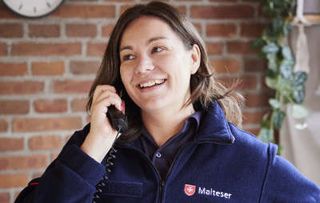 In einem Raum steht eine junge Frau mit einem Telefonhörer am Ohr. Sie trägt eine blaue Malteser-Jacke und lächelt. 