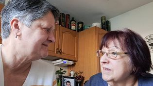 Zwei Frauen sitzen an einem Küchentisch mit Tassen, im Hintergrund Küchenschränke und Dekorationsflaschen sichtbar.