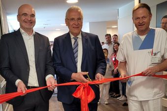 Drei Männer durchtrennen ein rotes Band bei einer Eröffnung, umgeben von weiteren Personen.